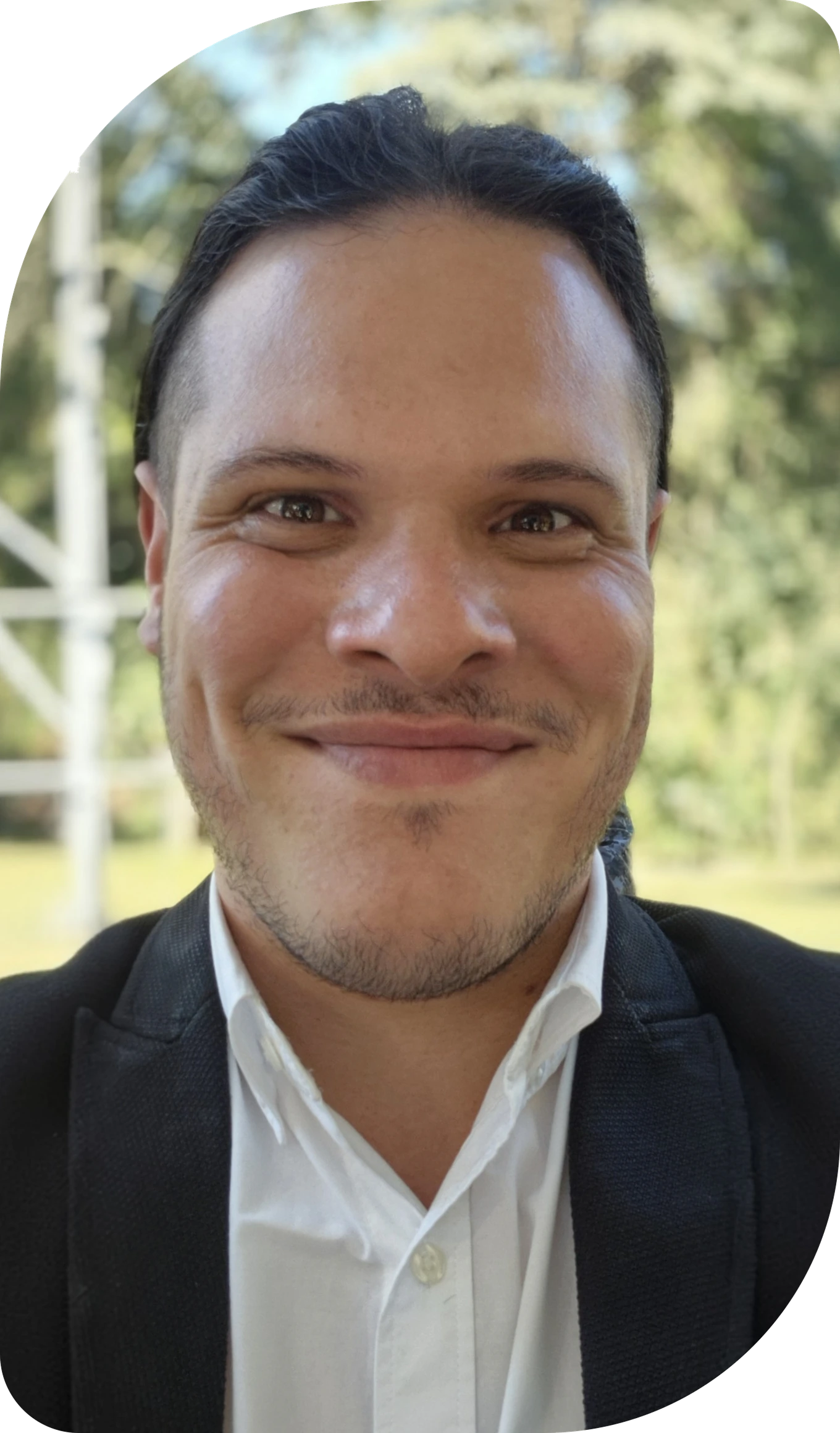 Foto de Érick Jesus, sorrindo, com blazer escuro sobre camisa branca, em ambiente externo com vegetação ao fundo
